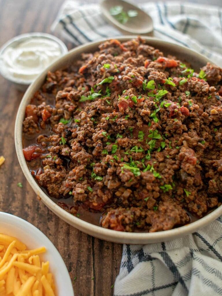 A bowl of ground beef garnished with chopped herbs, with shredded cheese and a bowl of sour cream nearby on a wooden table.