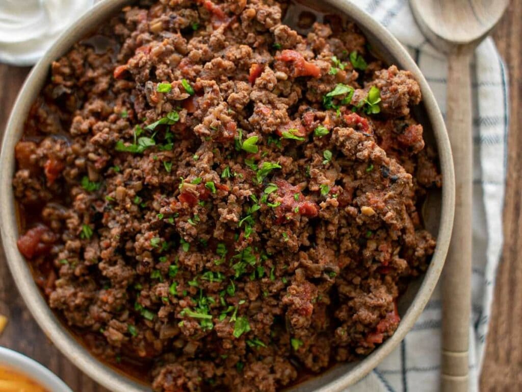 Cooked ground beef mixed with diced tomatoes and topped with chopped parsley, set on a cloth napkin with a wooden spoon nearby.