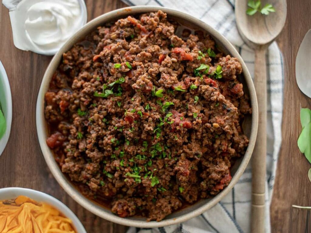 A bowl of cooked ground beef with herbs and tomato, surrounded by sour cream and fresh cilantro on a wooden table.