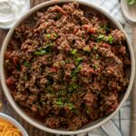 A bowl of ground beef garnished with chopped herbs, placed on a cloth napkin on a wooden table.