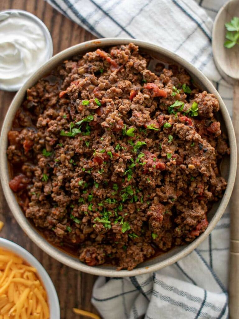 A bowl of ground beef garnished with chopped herbs, placed on a cloth napkin on a wooden table.