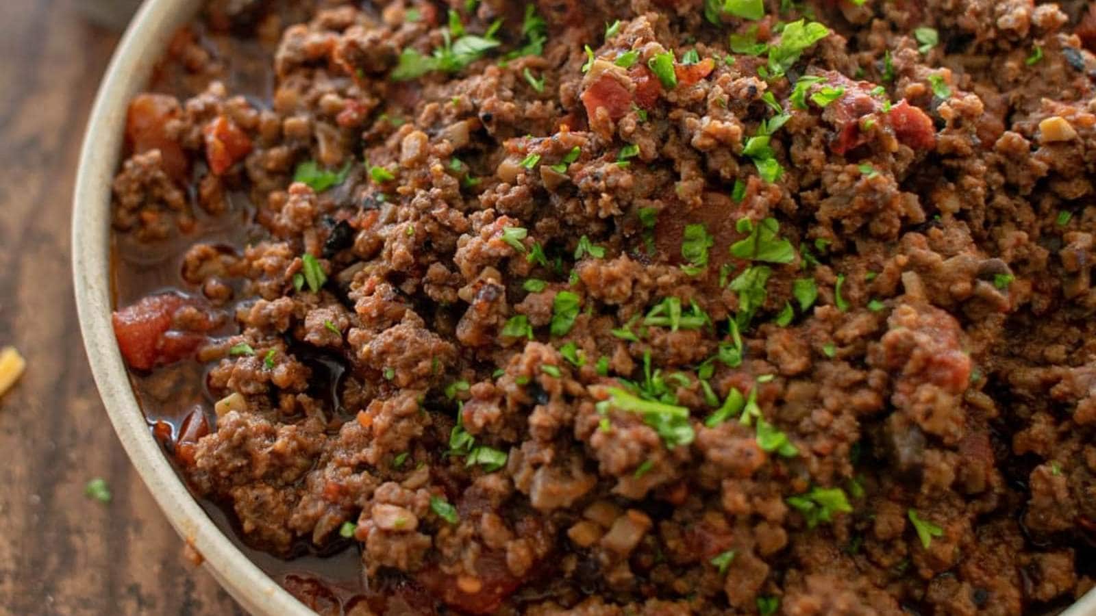 A close-up of a bowl filled with cooked ground beef mixed with diced tomatoes and garnished with chopped parsley.
