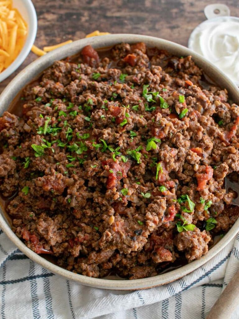A bowl of cooked ground beef with tomatoes, topped with chopped herbs, sits on a cloth napkin.