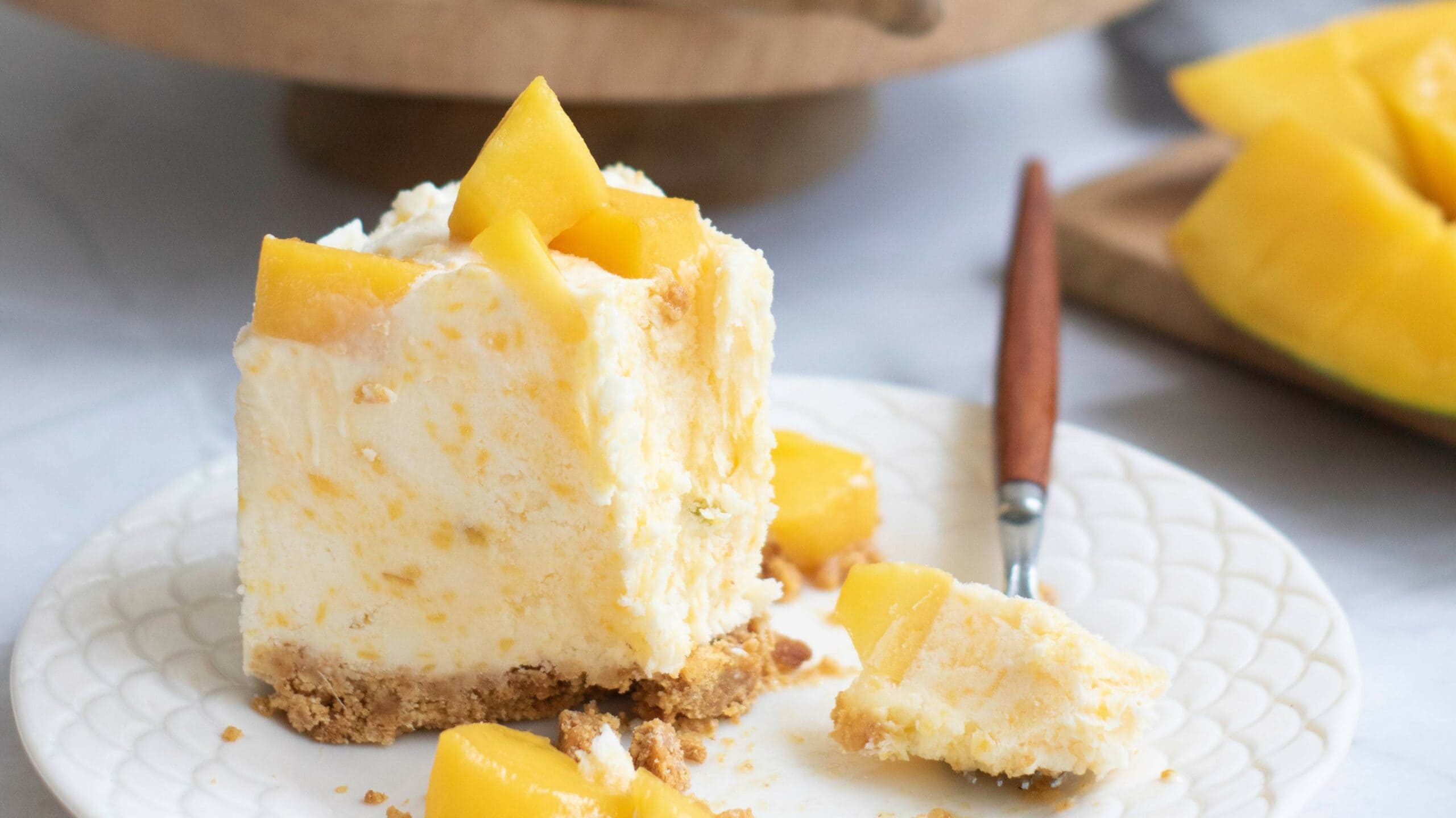 A slice of mango cheesecake with a crumbly crust, topped with chunks of fresh mango, sits on a white plate with a fork.