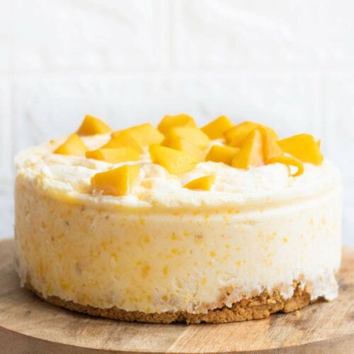 A round cheesecake with a crumbly crust, topped with pieces of fresh yellow mango, displayed on a wooden cake stand.