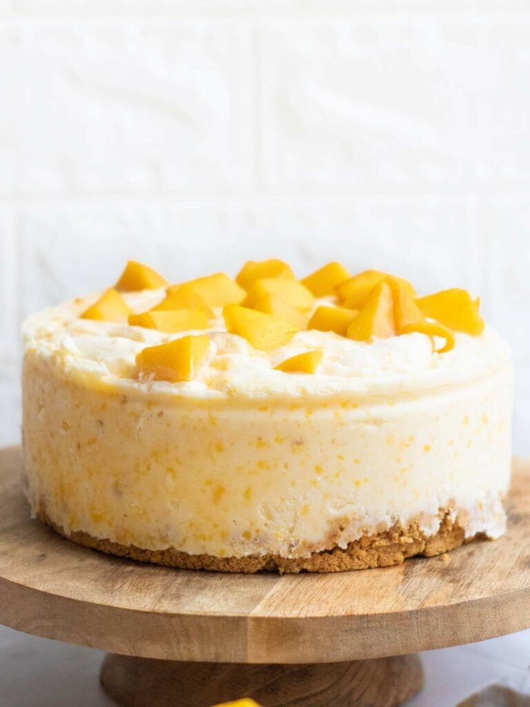 A round cheesecake with a crumbly crust, topped with pieces of fresh yellow mango, displayed on a wooden cake stand.