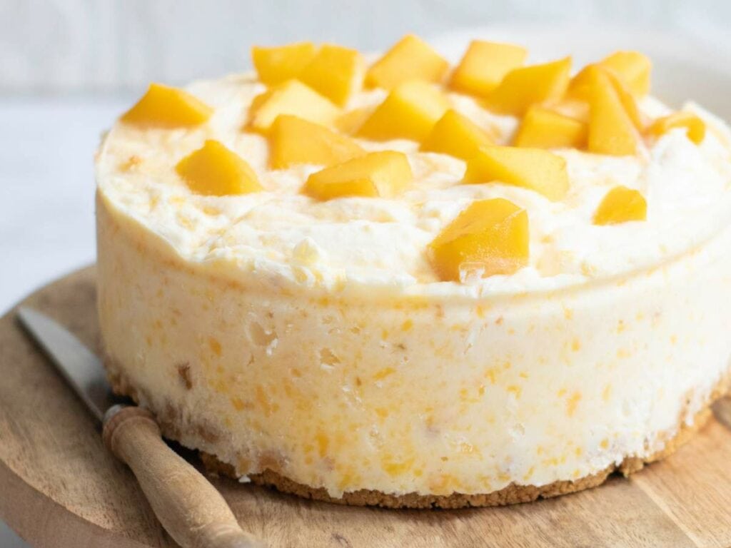 A round creamy cake topped with diced mango pieces sits on a wooden board next to a knife.