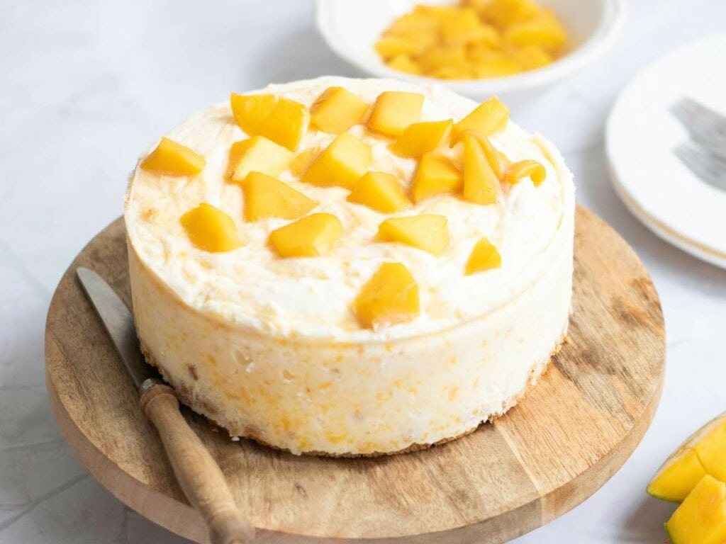 A round cheesecake topped with diced mango sits on a wooden board next to a knife, with a bowl of mango pieces and a plate in the background.