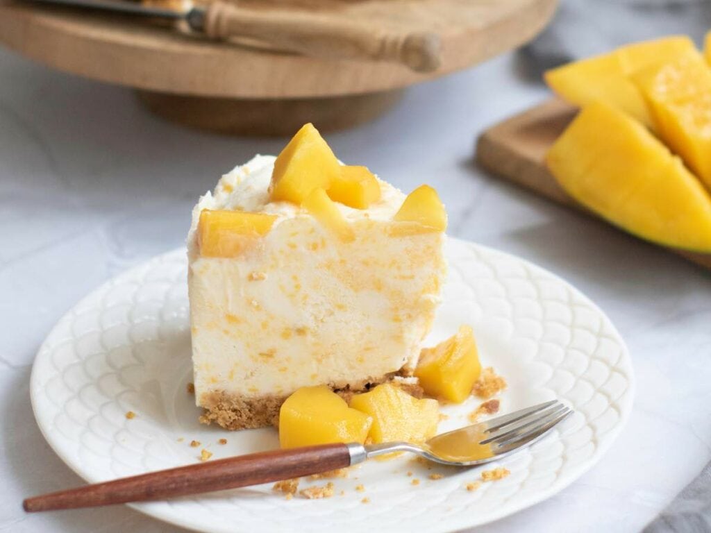 A slice of cheesecake with chunks of mango on top and beside it, served on a white plate with a fork.