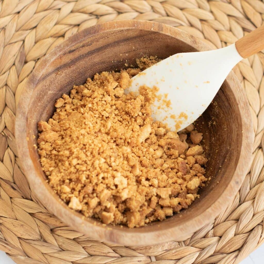 A wooden bowl filled with crushed graham crackers and a white spatula, placed on a woven placemat.