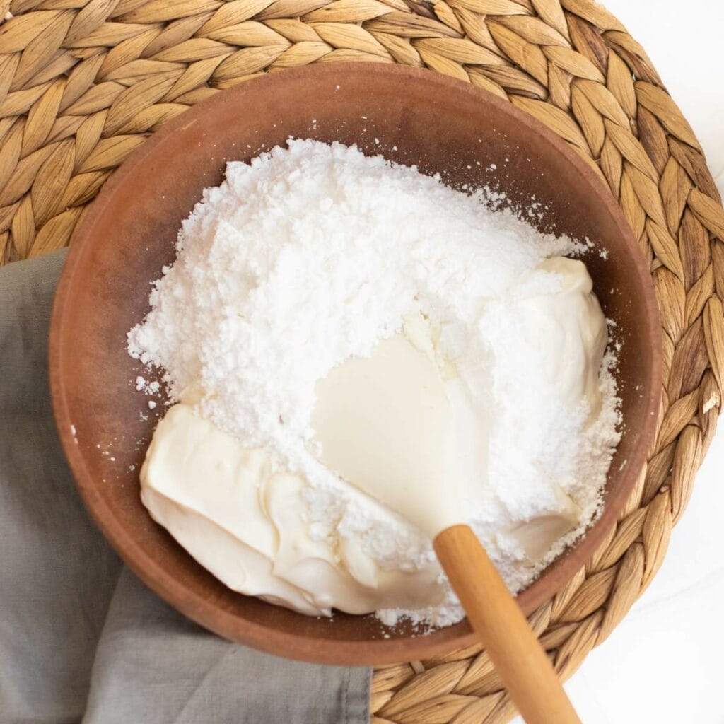 A wooden bowl containing cream cheese and powdered sugar being mixed with a white spatula.