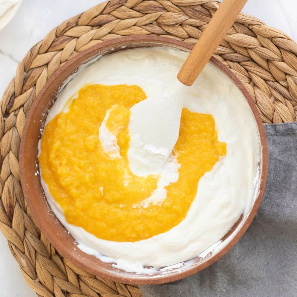 A wooden bowl with whipped cream and a layer of mango puree being mixed with a white spatula.