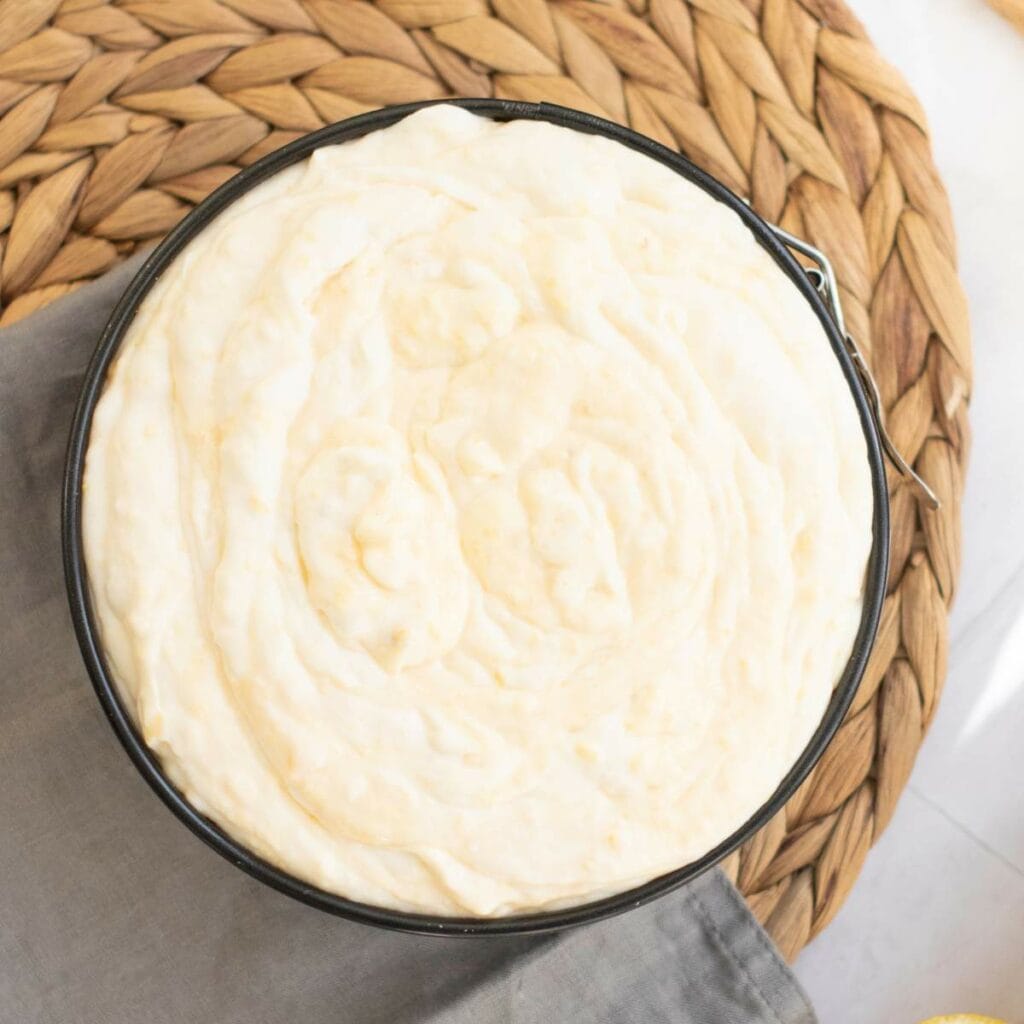 A round baking pan filled with uncooked cheesecake batter sits on a woven placemat with a gray napkin underneath.