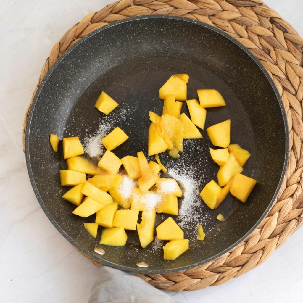 Chopped mango and granulated sugar in a nonstick frying pan on a woven placemat.