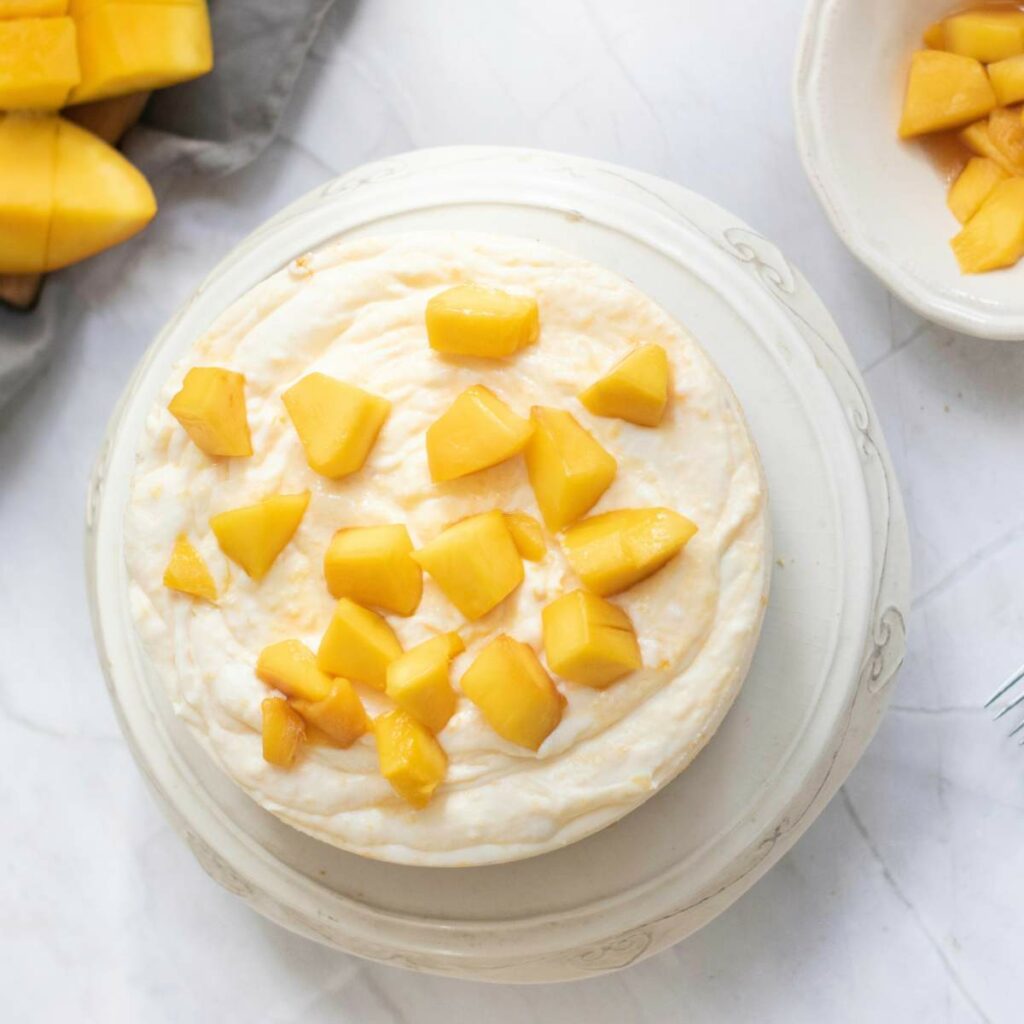 A round cheesecake topped with diced mango pieces, placed on a white plate next to a bowl and board with more chopped mango.
