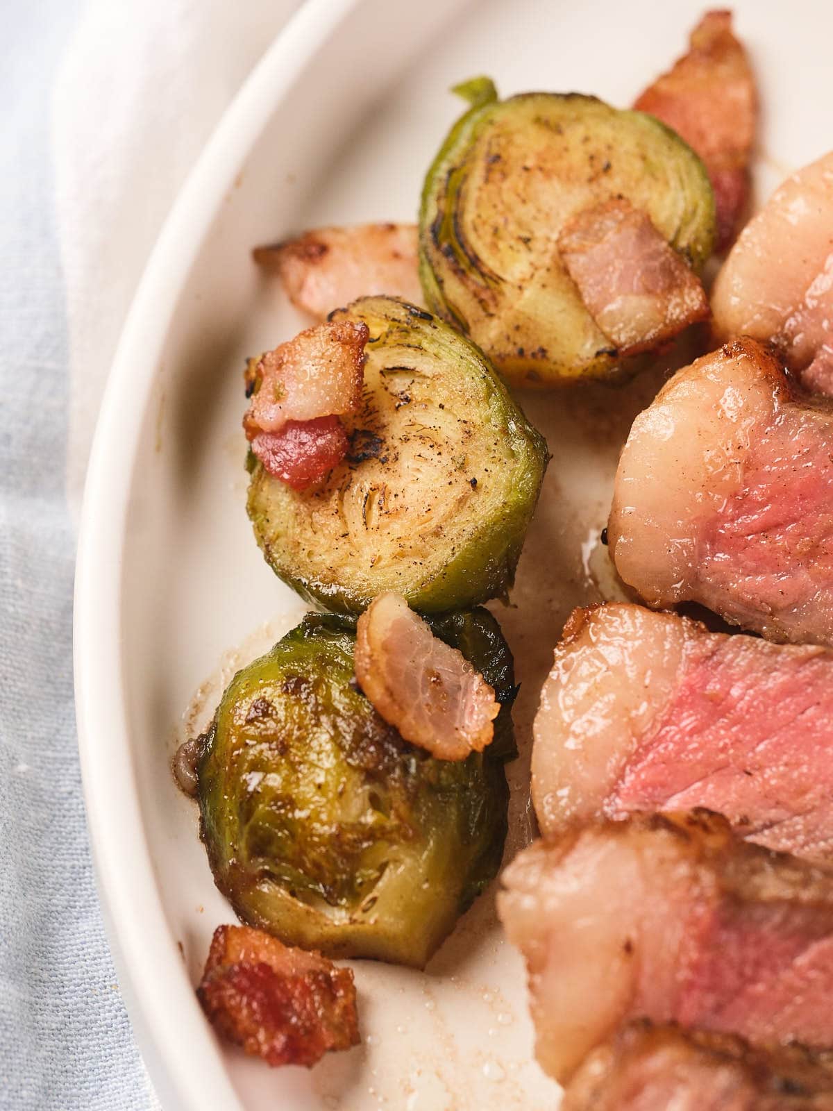 Close-up of three cooked Brussels sprouts and bacon pieces on a white plate next to slices of medium-rare steak.