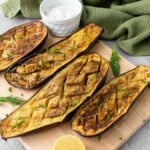 Four cooked eggplant halves with a crisscross pattern on a wooden board.