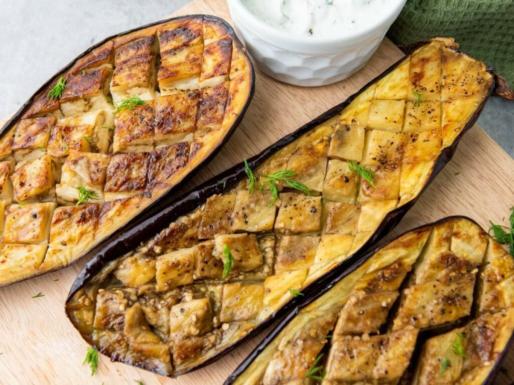 Three cooked eggplant halves with scored tops are served on a wooden board, garnished with herbs.