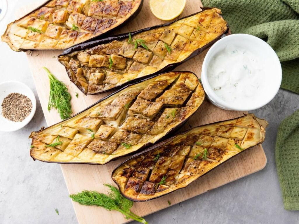 Four seasoned eggplant halves on a wooden board, scored in a diamond pattern, served with a bowl of white sauce.