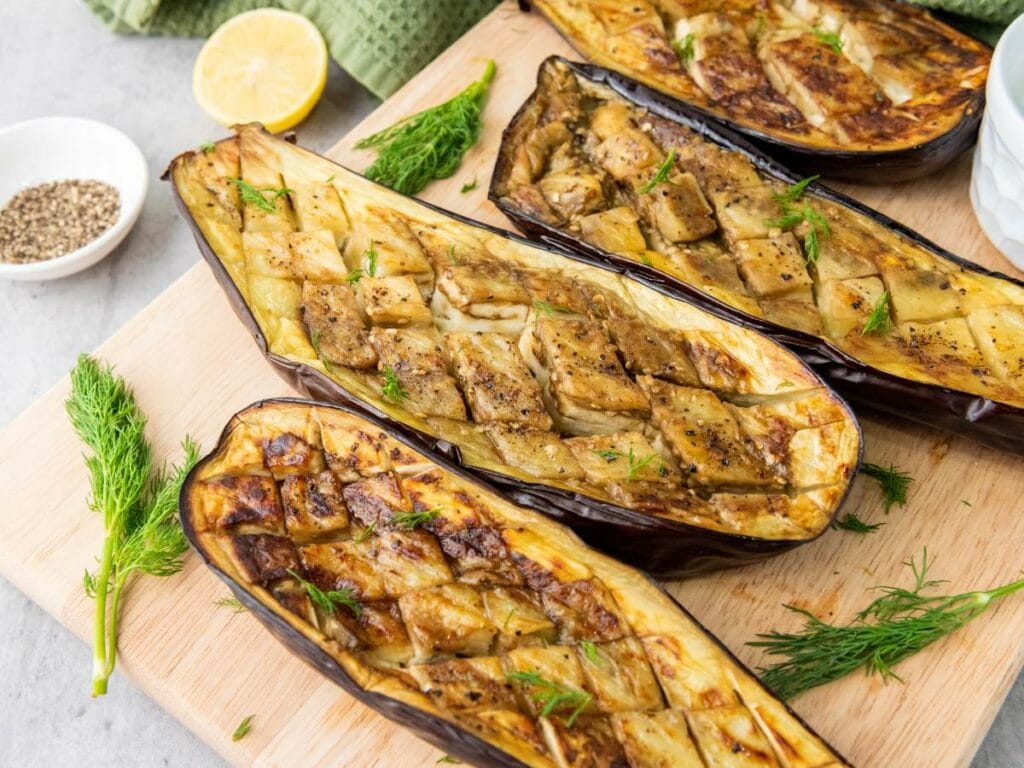 Four cooked eggplant halves with scored tops are arranged on a wooden board, garnished with fresh dill.