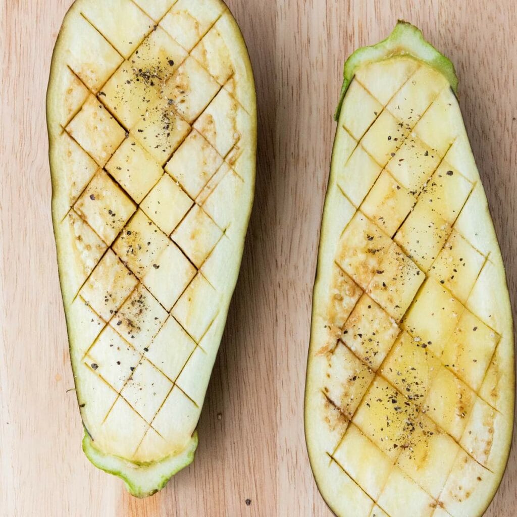 Two eggplant halves scored with a crosshatch pattern, seasoned with salt and pepper.