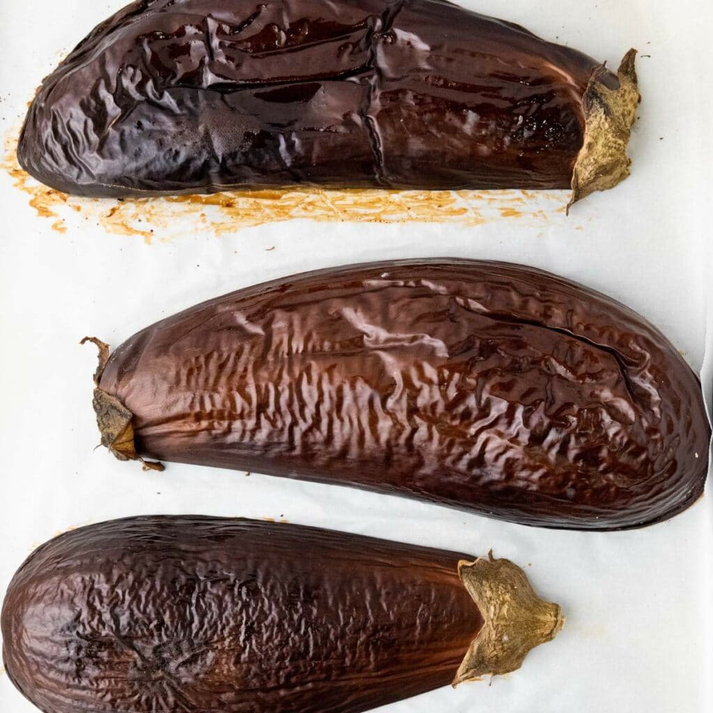 Three halves roasted eggplants with wrinkled, browned skin are arranged upside down on a white surface.