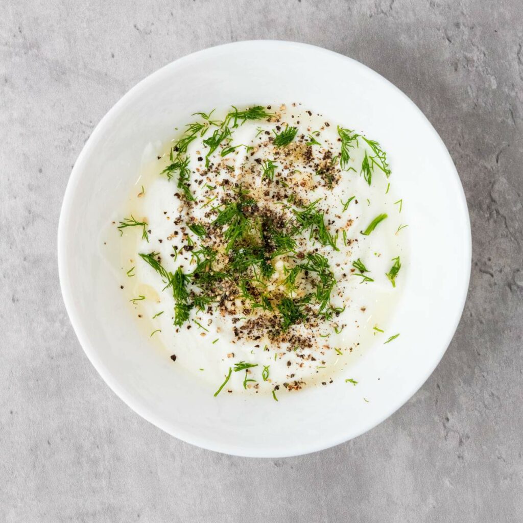 A white bowl filled with yogurt, garnished with fresh dill, black pepper, and a drizzle of olive oil.