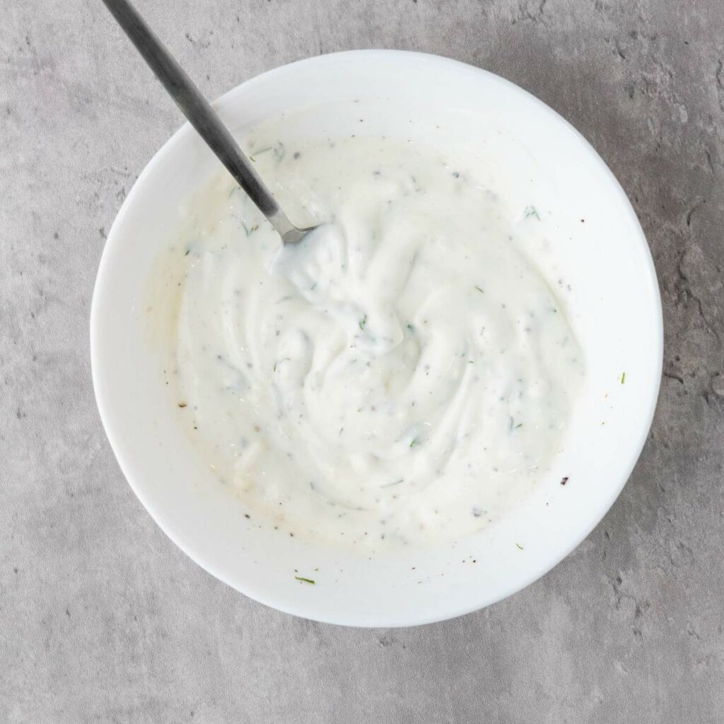 A white bowl filled with creamy white sauce or dip, speckled with herbs and black pepper, with a metal spoon inside.