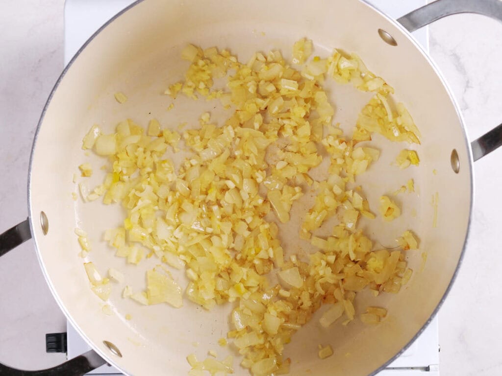Chopped onions sautéing in a light-colored pan on a stovetop.
