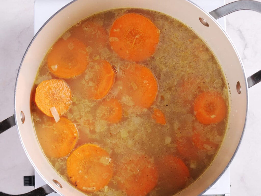 A pot of soup with sliced carrots, chopped onions, and broth simmering on a stove.
