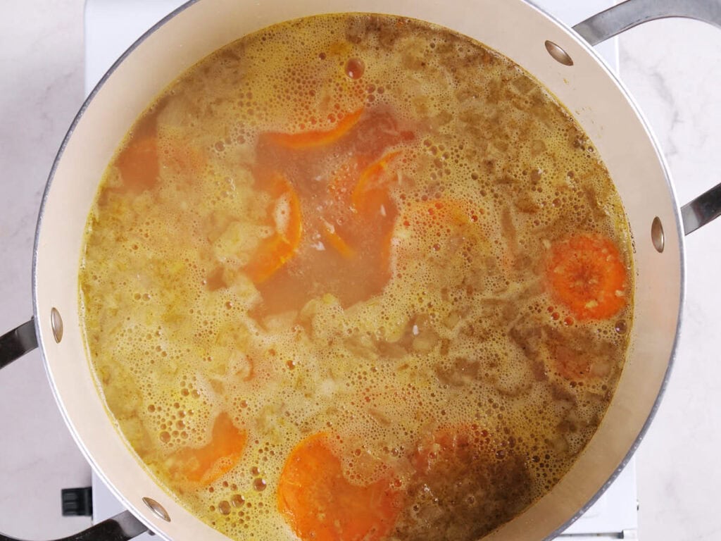 A pot of soup with visible sliced of carrots simmering in broth on a stovetop.