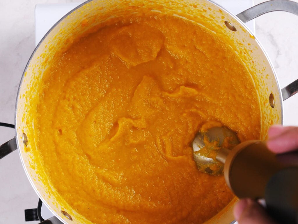 A hand uses an immersion blender to puree orange soup in a large metal pot on a stovetop.