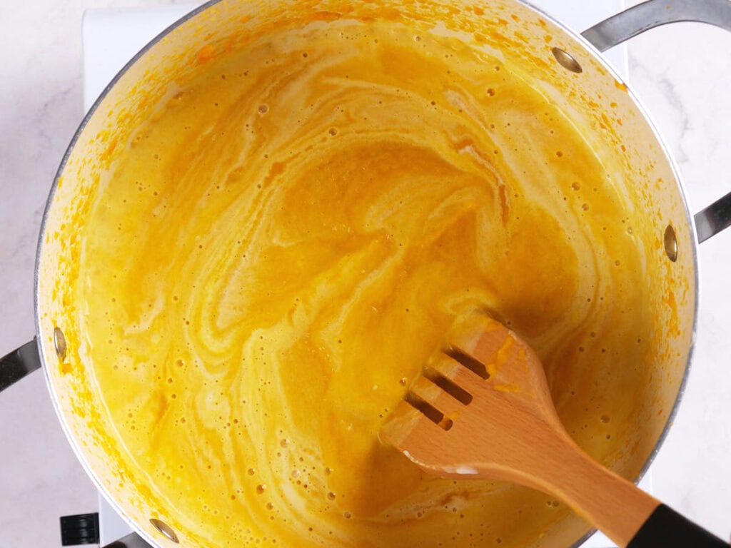 A pot of yellow liquid being stirred with a wooden spatula, showing swirls on the surface.
