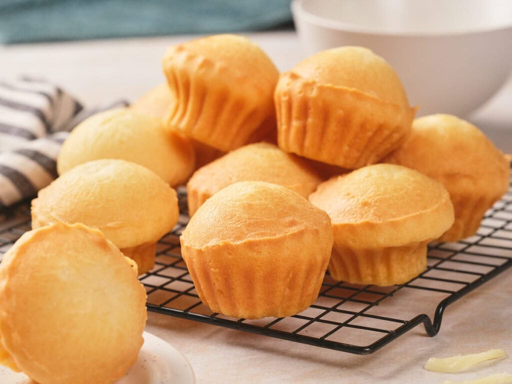 A group of plain golden muffins cooling on a black wire rack, with a bowl and striped towel in the background.