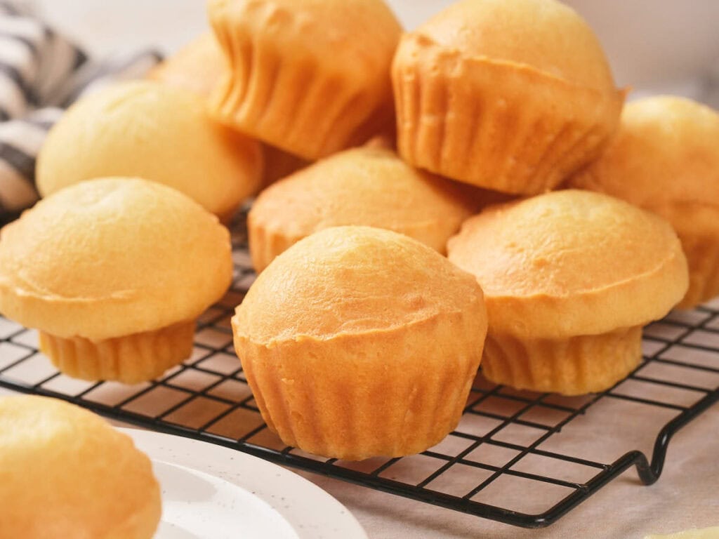 A batch of plain, golden brown muffins cooling on a black wire rack, with a white plate partly visible in the foreground.