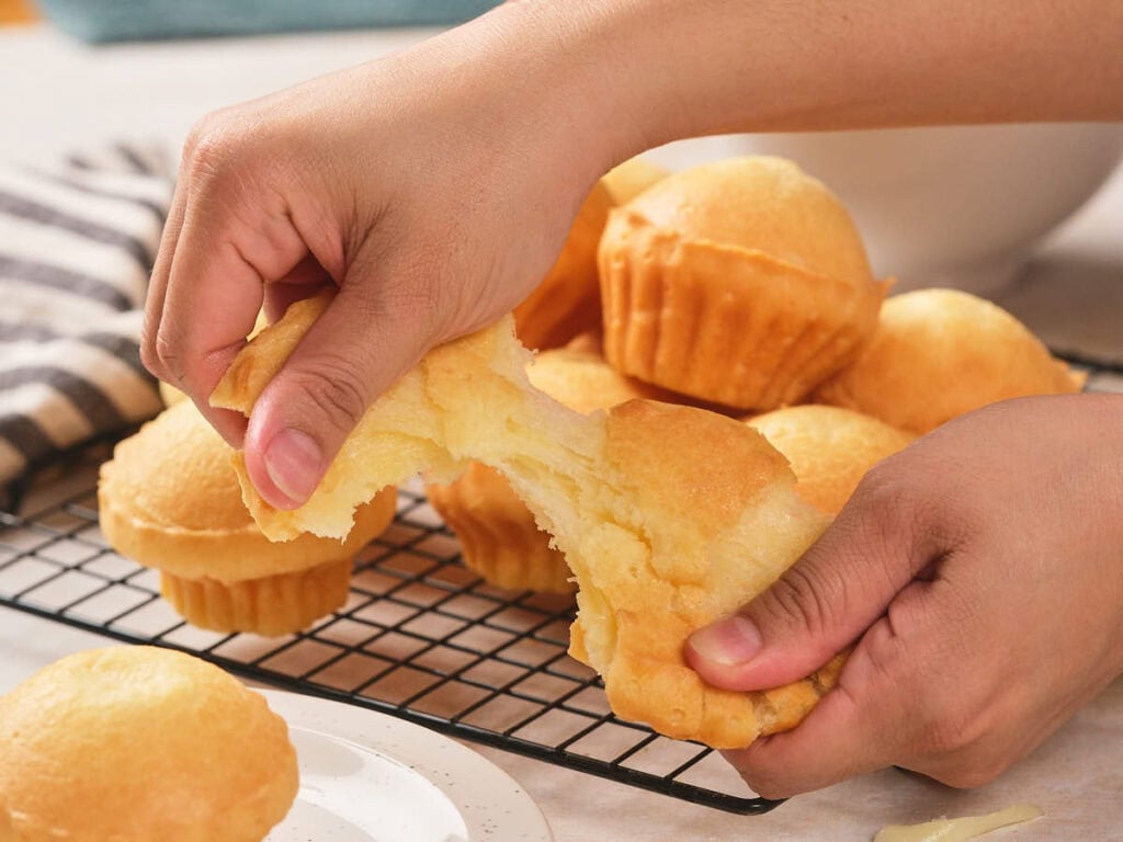 A person pulls apart a muffin, showing its soft, fluffy interior, with several muffins cooling on a wire rack.