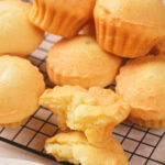 A group of golden-brown muffins are stacked on a cooling rack, with one muffin broken in half showing its soft, fluffy interior.