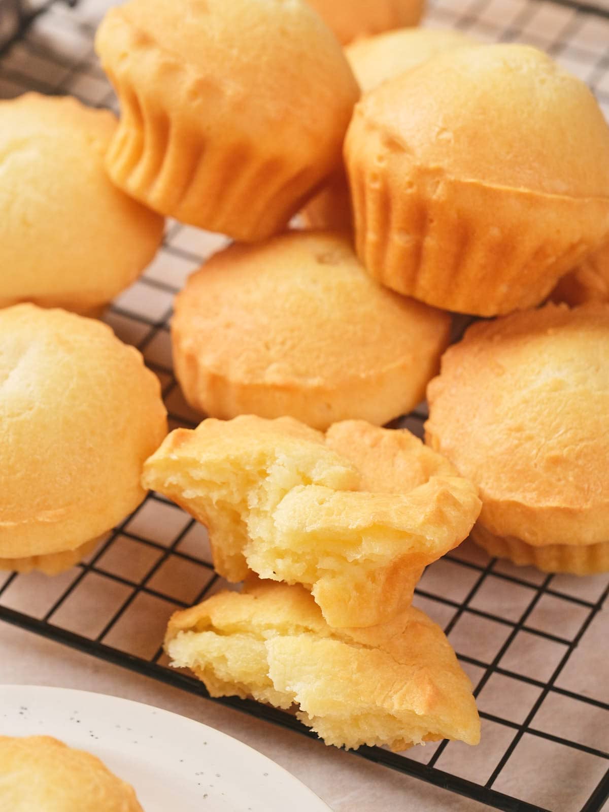 A group of golden-brown muffins are stacked on a cooling rack, with one muffin broken in half showing its soft, fluffy interior.