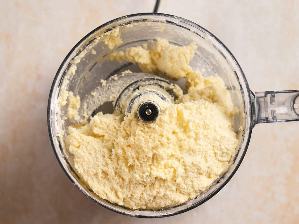 Overhead view of a food processor containing a coarse, pale-yellow mixture, likely butter or dough.