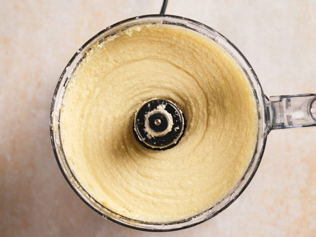 A food processor filled with a smooth, pale mixture viewed from above on a light-colored surface.