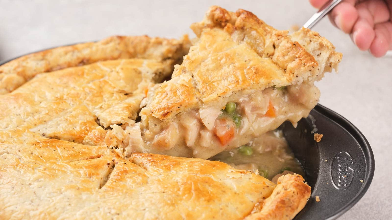 A hand lifts a slice of chicken pie from a baking dish, showing a golden crust and creamy filling with vegetables.