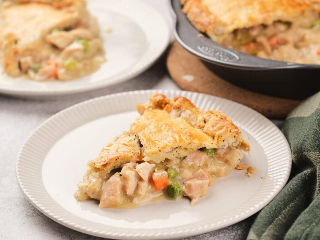 A slice of chicken pie with vegetables on a white plate, with the remaining pie in a baking pan in the background.
