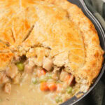 A close-up of a baked chicken pie in a pan with a golden crust, partially sliced to show the creamy filling.