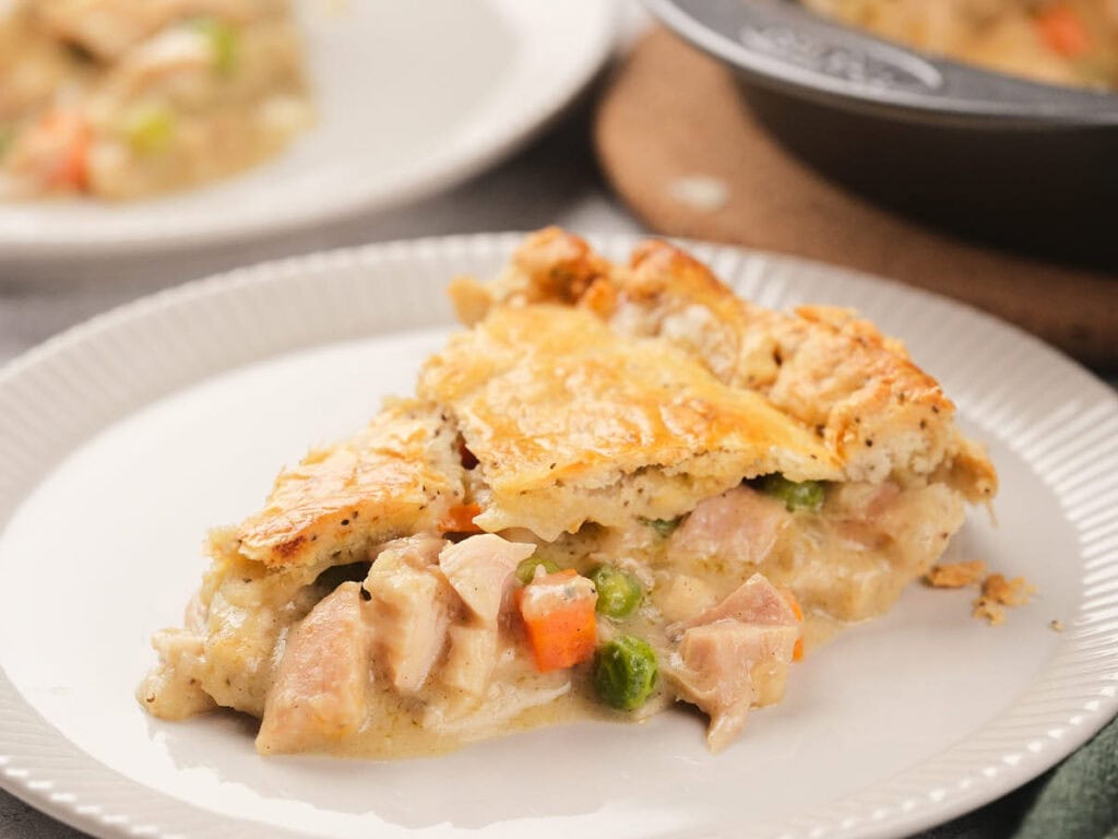 A slice of chicken pie with a golden-brown crust, filled with chicken, peas, carrots, and creamy sauce on a white plate.