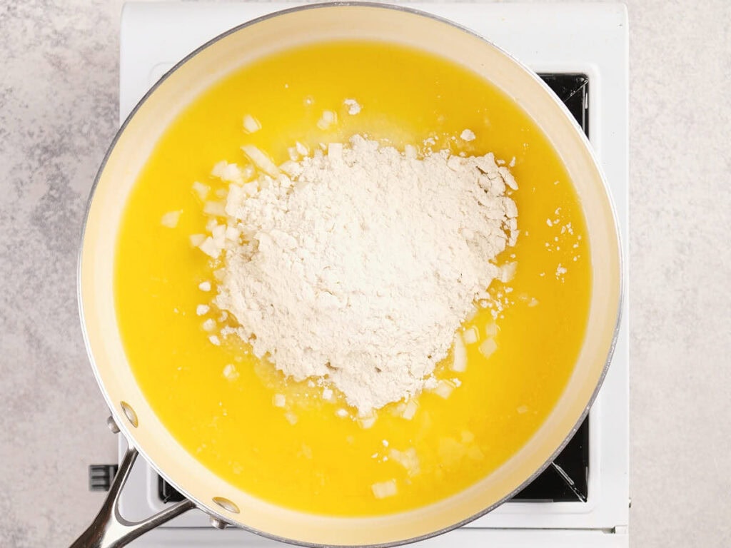 A pan on a stovetop contains melted butter, chopped onions, and a mound of flour.