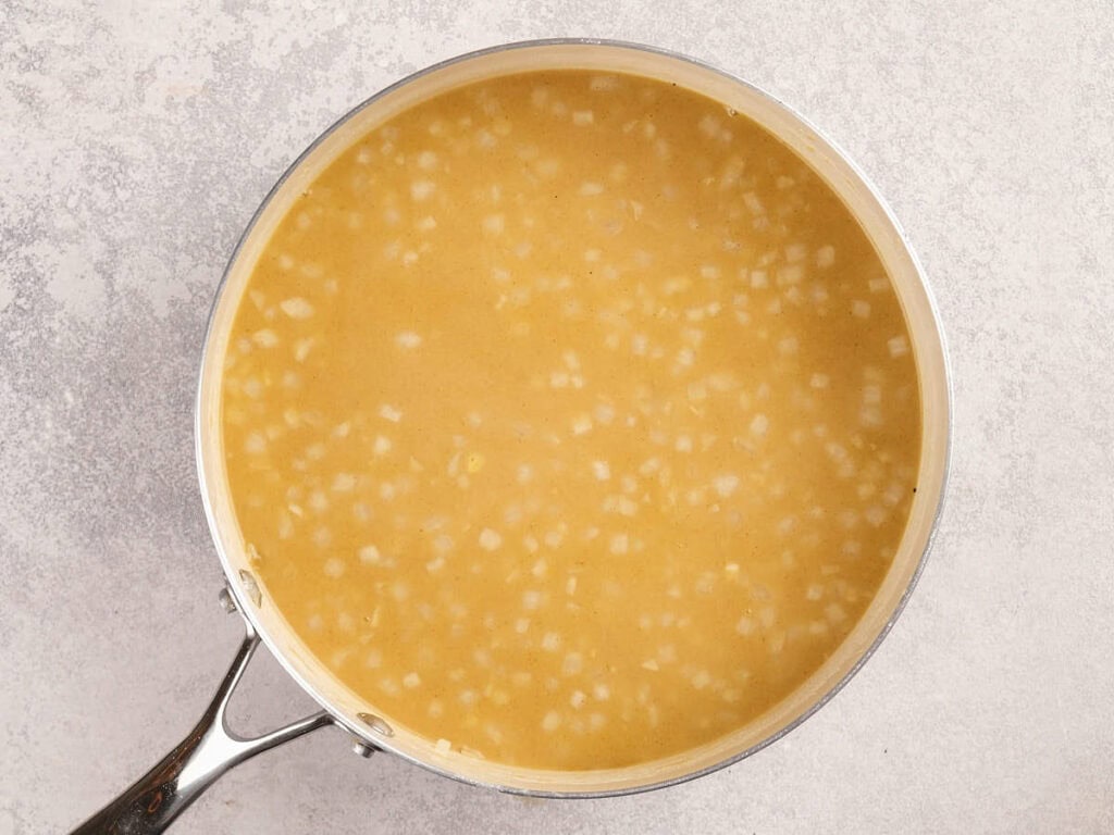 A saucepan filled with a yellow liquid containing small diced onions, viewed from above on a light-colored surface.