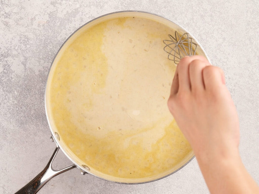 A hand whisking a light yellow sauce in a stainless steel pan on a light-colored countertop.