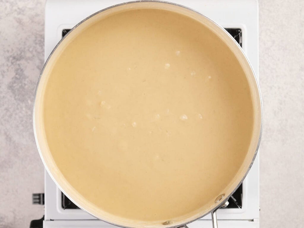 A stainless steel saucepan filled with creamy beige sauce is heating on a white stovetop.