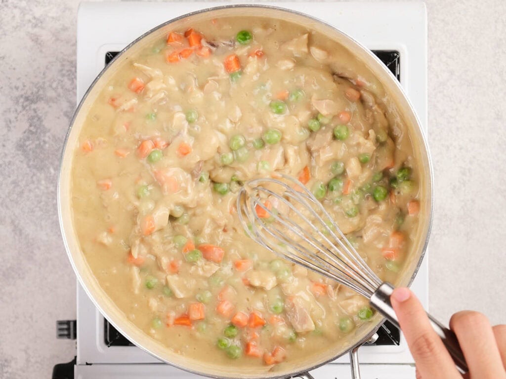 A hand uses a whisk to stir a thick mixture of peas, carrots, and sauce in a large pan on a stovetop.