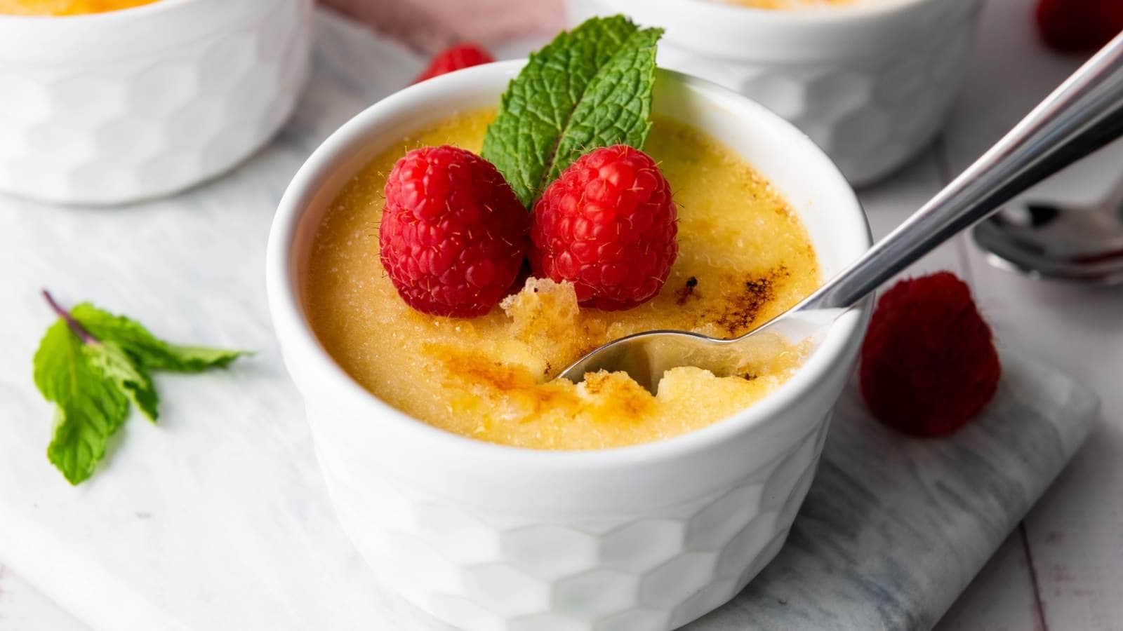 A white ramekin filled with crème brûlée, topped with two raspberries and a mint leaf, with a spoon digging into the dessert.